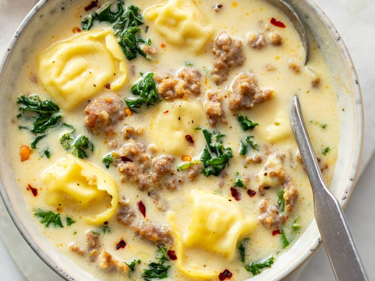 Close-up of a bowl of creamy sausage tortellini soup with kale and red pepper flakes, spoon on the side